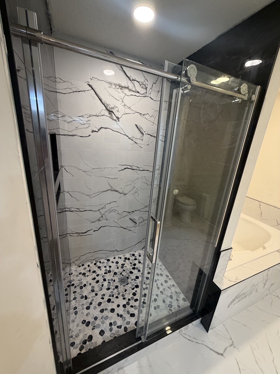 Walk-in shower with marble-look wall tile, pebble floor, and glass door